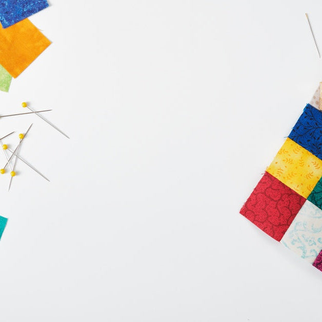 Quilting supplies on a white surface. Perfectly cut squares, pins, scissors, a rotary cutter, and more are laid out neatly.