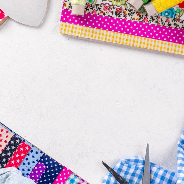 Quilting supplies arranged on a white surface. Visible are fabric squares, scissors, thread, a ruler, and a rotary cutter.