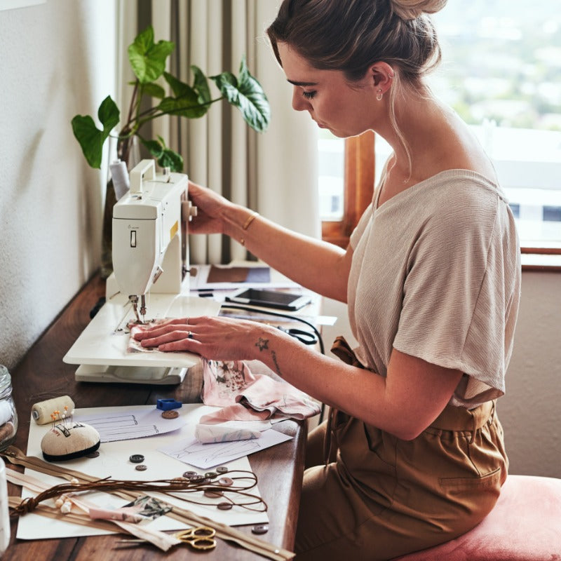A woman using a sewing machine on a desk strewn with notions. She sits on a pink-cushioned stool by a window.