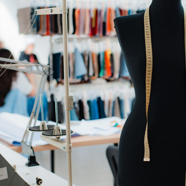 A black dress form with a measuring tape draped over its shoulders in a sewing studio with machines and fabric samples behind it.