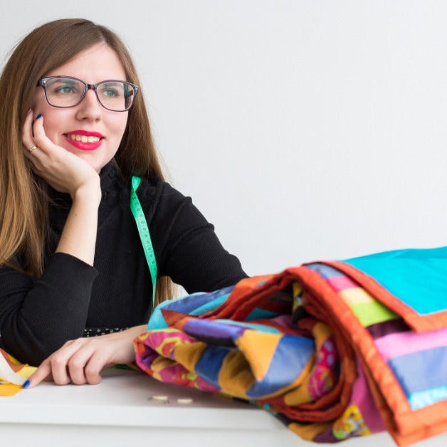 A woman with glasses smiling as she sits at a table behind a colorful folded quilt and a sewing machine.