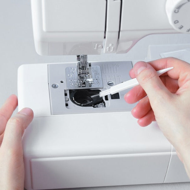A close-up of hands using a small brush to clean lint from the bobbin area of a white sewing machine.