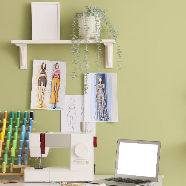 A sewing workspace with a machine, a colorful thread rack, an open laptop, and fashion sketches on a green wall.