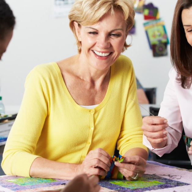 Three smiling women gathered around a colorful quilt laid across a crafting table in a shared studio space.