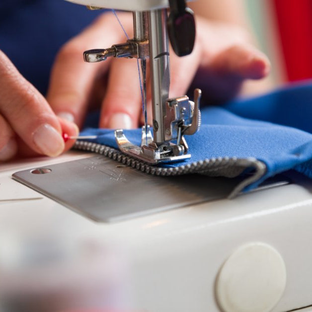 A close-up of two hands sewing on a zipper to a blue piece of fabric while removing a pin next to the foot.