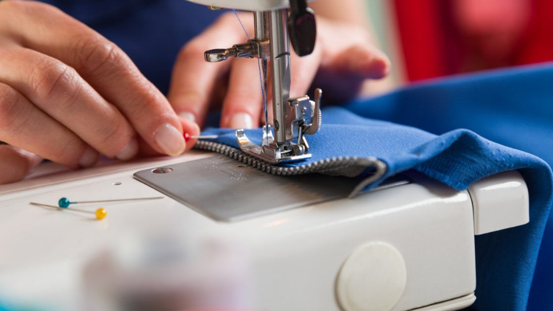 A close-up of two hands sewing on a zipper to a blue piece of fabric while removing a pin next to the foot.