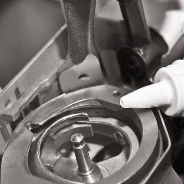 A close-up of a white bottle next to the bobbin holder of a sewing machine with multiple screws and moving parts.