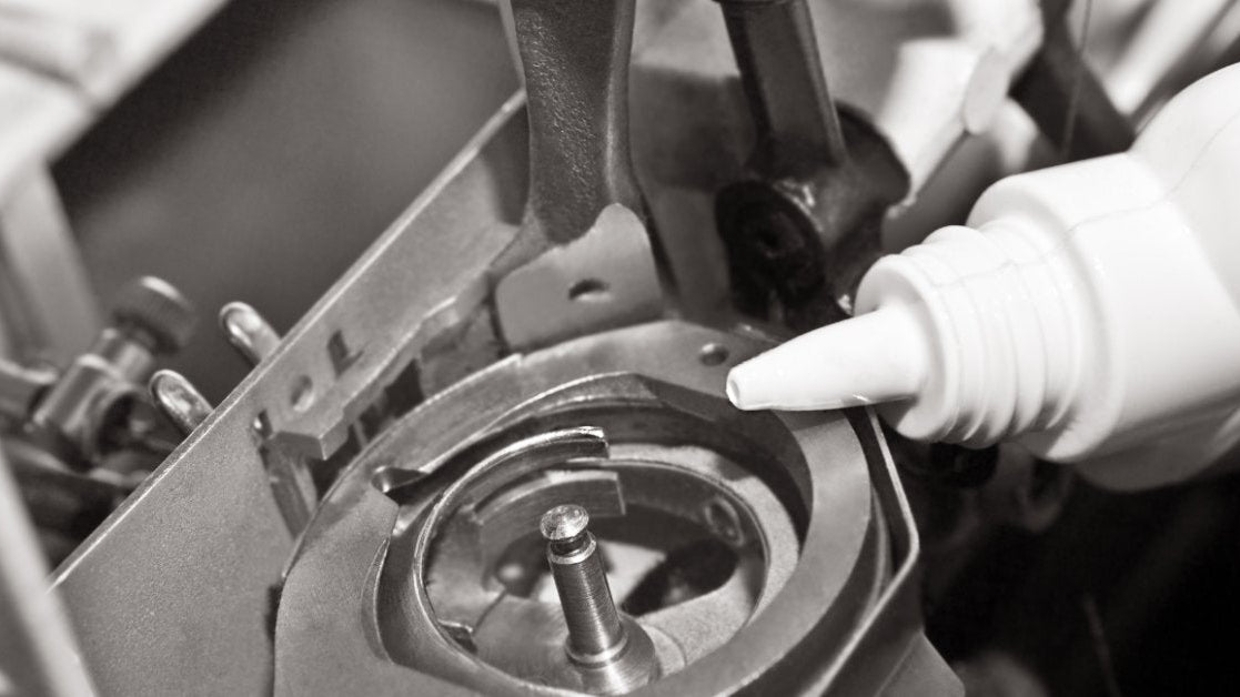 A close-up of a white bottle next to the bobbin holder of a sewing machine with multiple screws and moving parts.