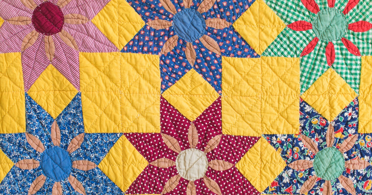 A close-up of a quilt.The primary color is yellow, and the flower designs are made with neatly cut, colorful, patterned fabrics.