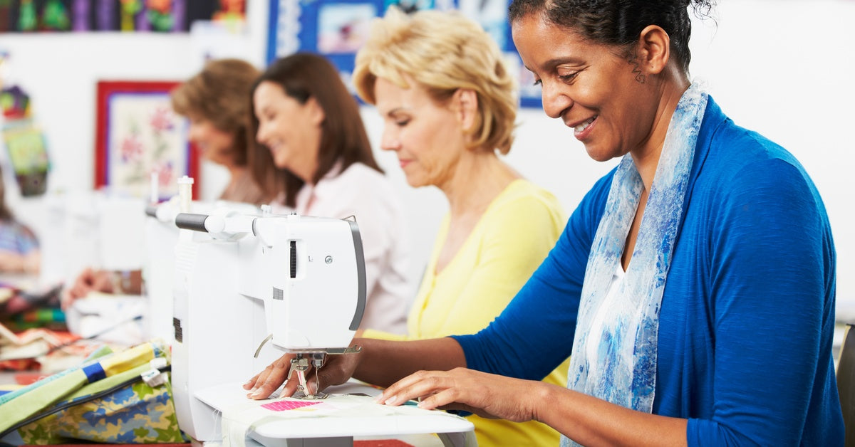 Four women in a sewing class. The sit in a row at a single table, each with their own sewing machine.