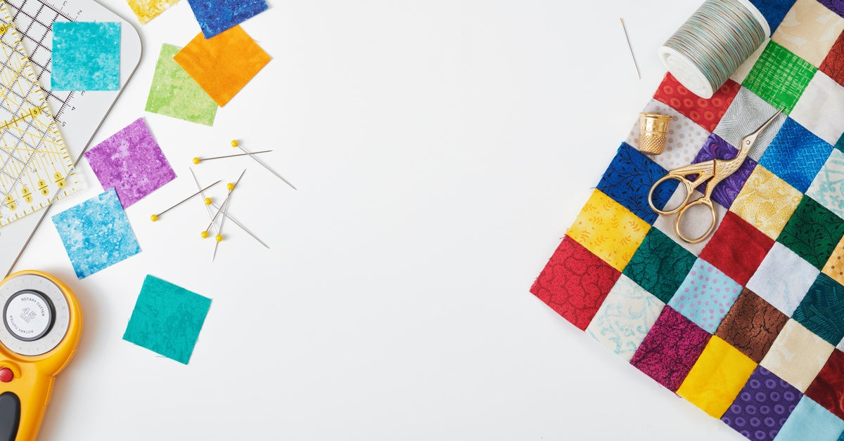 Quilting supplies on a white surface. Perfectly cut squares, pins, scissors, a rotary cutter, and more are laid out neatly.