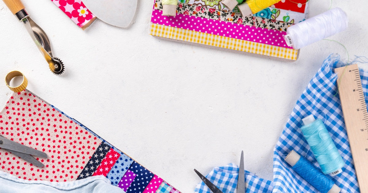 Quilting supplies arranged on a white surface. Visible are fabric squares, scissors, thread, a ruler, and a rotary cutter.