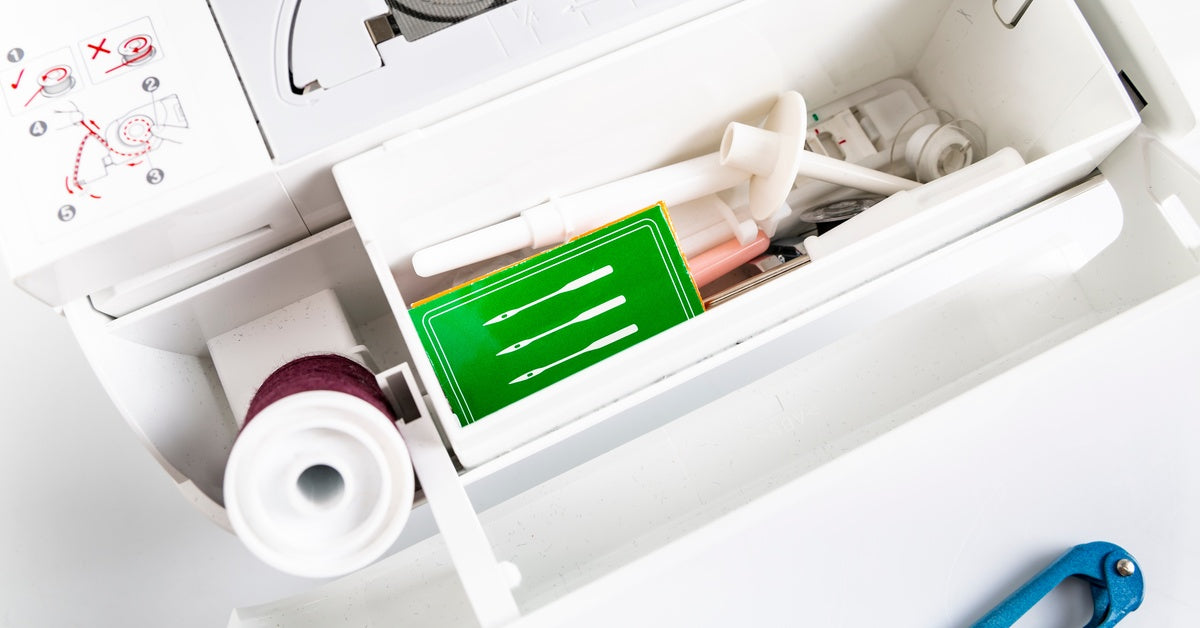 The open accessories compartment of a white sewing machine, revealing needles, thread, bobbins, presser feet, and more.