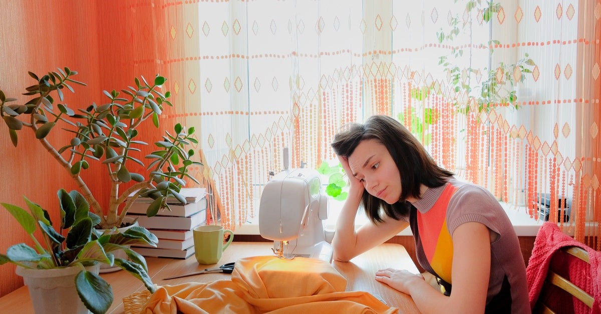 A tired, frustrated woman sitting at a table by a window and looking at her sewing machine mid-project.