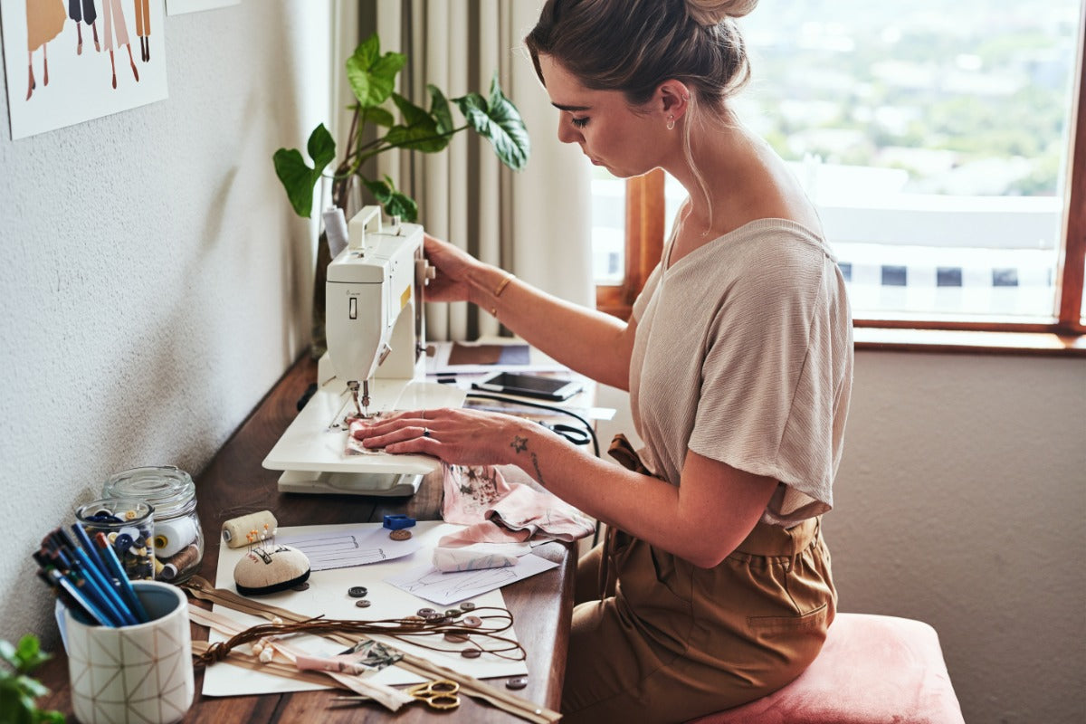 A woman using a sewing machine on a desk strewn with notions. She sits on a pink-cushioned stool by a window.