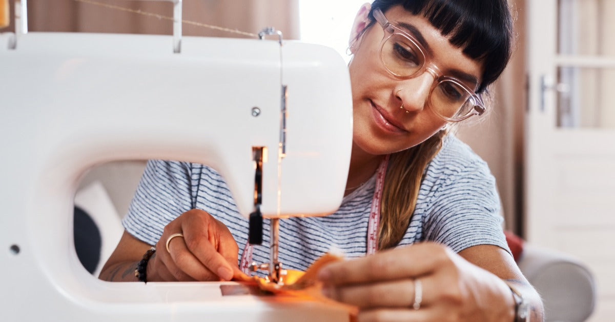 A woman in glasses guides orange fabric under a white sewing machine needle. She's inside her home.