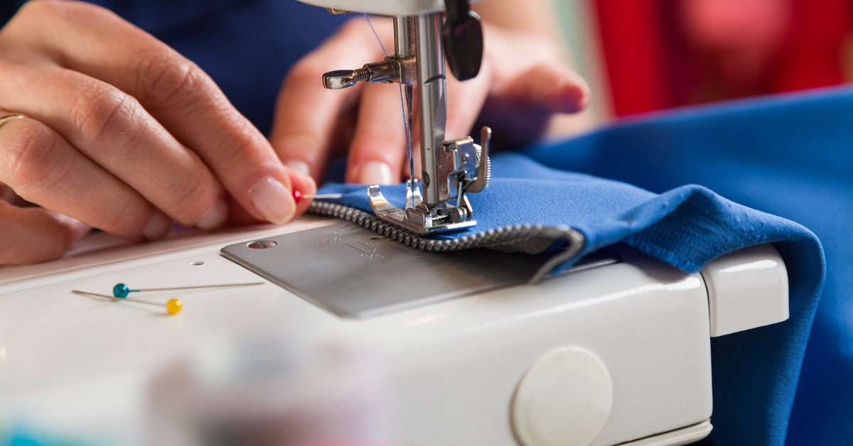 A close-up of hands guiding blue fabric and a zipper under a sewing machine needle while placing a pin below the foot.