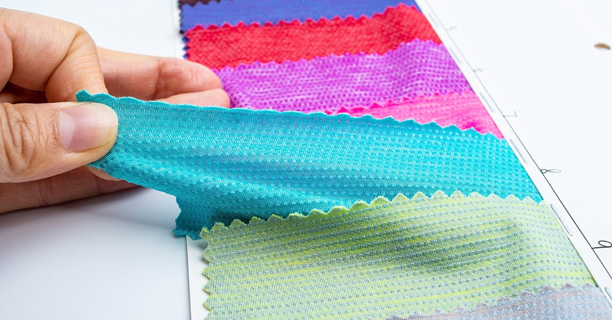 A hand lifts and pulls a stretchy teal fabric swatch from a sample strip, next to the same fabrics in different bright colors.
