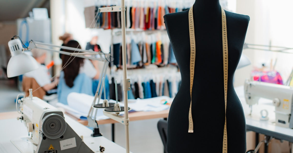 A black dress form with a measuring tape draped over its shoulders in a sewing studio with machines and fabric samples behind it.