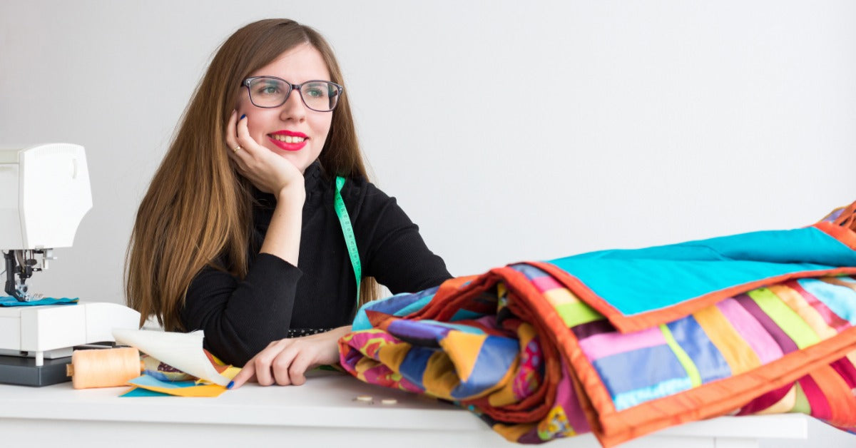 A woman with glasses smiling as she sits at a table behind a colorful folded quilt and a sewing machine.