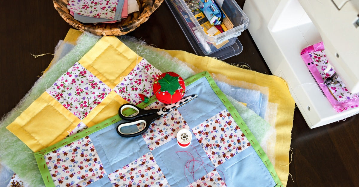 Several patchwork quilt squares, batting, scissors, a pincushion, thread, extra fabric squares, and a sewing machine on a table.