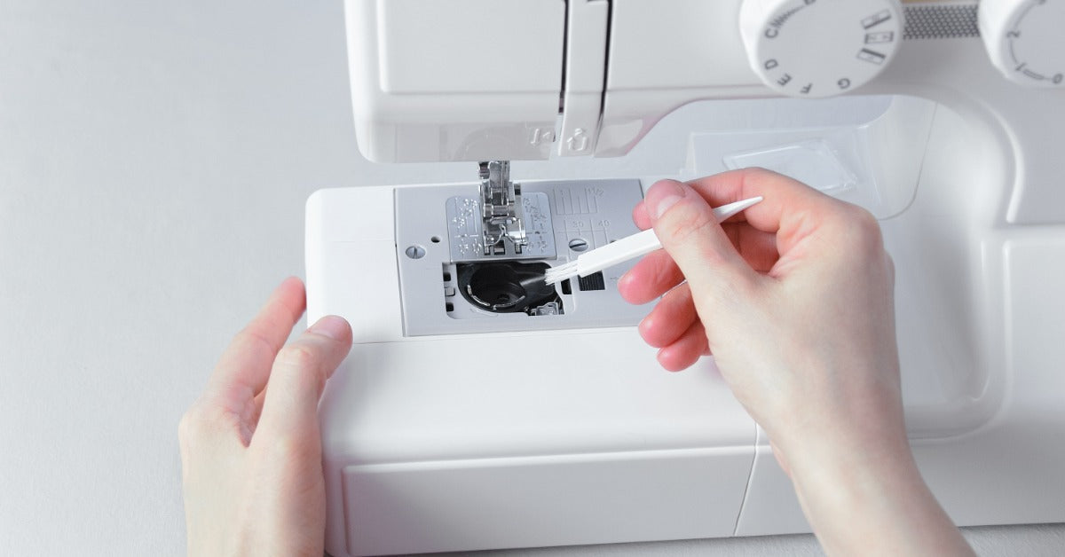 A close-up of hands using a small brush to clean lint from the bobbin area of a white sewing machine.