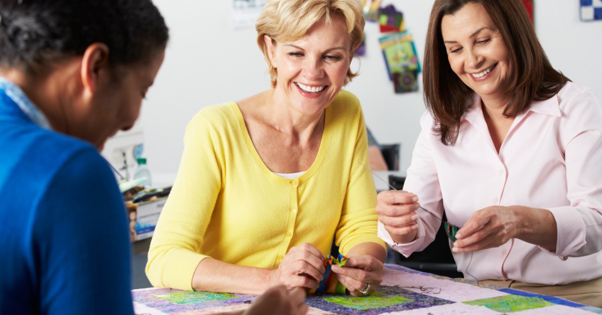 Three smiling women gathered around a colorful quilt laid across a crafting table in a shared studio space.
