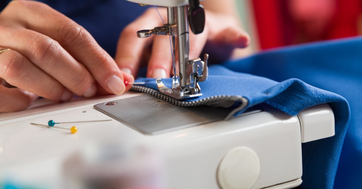 A close-up of two hands sewing on a zipper to a blue piece of fabric while removing a pin next to the foot.