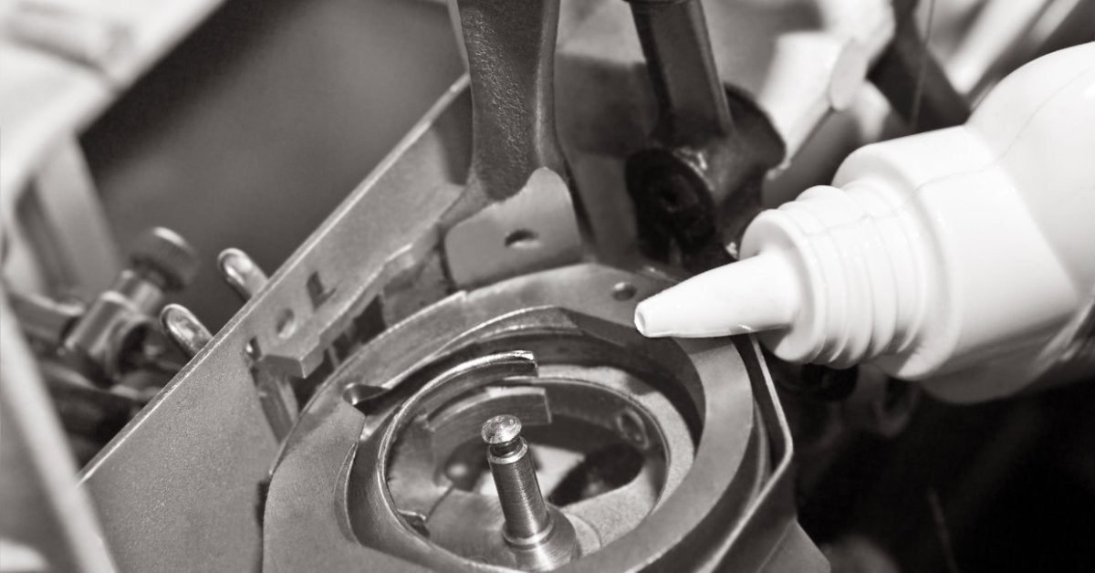 A close-up of a white bottle next to the bobbin holder of a sewing machine with multiple screws and moving parts.